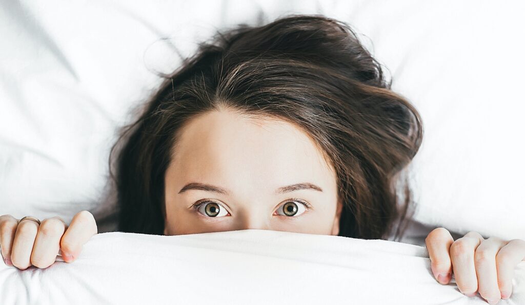 Photo of a young woman's wide-eyed face, partly obscured by bedcovers pulled up over her nose.